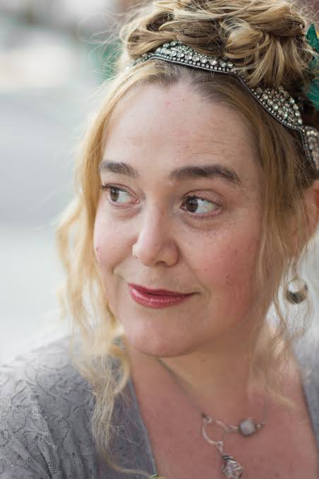 Portrait d'une femme avec des cheveux blonds ondulés, portant un bijou de tête scintillant et un haut gris. Elle regarde légèrement sur le côté avec une expression douce.