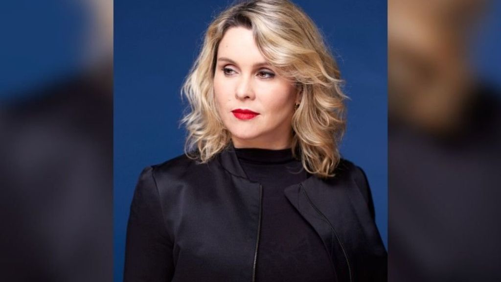 Portrait d'une femme avec des cheveux blonds bouclés et un rouge à lèvres rouge, portant une veste noire sur fond bleu.