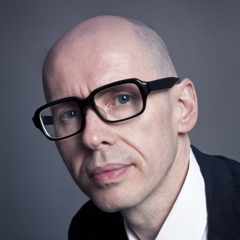 Portrait d'un homme chauve portant des lunettes noires, avec un regard sérieux et un fond neutre.